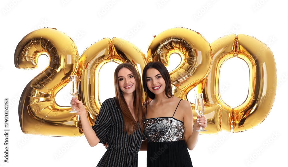 Happy young women with glasses of champagne near golden 2020 balloons on white background. New Year celebration