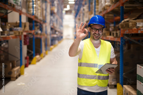 Successful goods distribution and warehouse organization. Warehouse worker standing in large storage center and showing OK hand gesture satisfied on delivering goods.