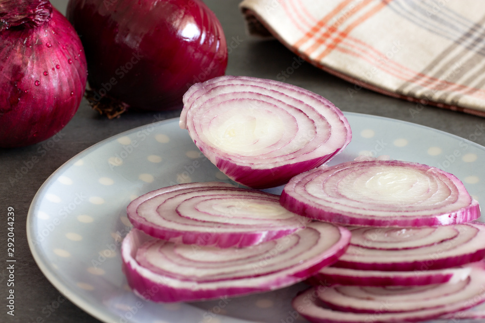 close up of red onion slices on blue plate with onions in the background on gray surface close up of red onion slices on blue plate with onions in the background on gray surface