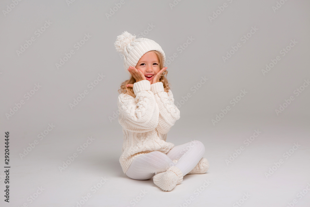 Winter clothes. Portrait of a little curly-haired girl in a knitted white winter hat. little blonde girl in white knitted hat and sweater smiling light background isolate, space for text Winter clothes. Portrait of a little curly-haired girl in a knitted white winter hat. little blonde girl in white knitted hat and sweater smiling light background isolate, space for text