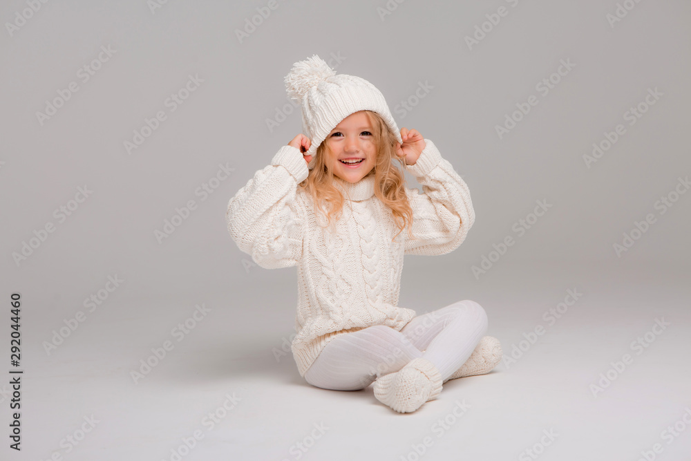 Winter clothes. Portrait of a little curly-haired girl in a knitted white winter hat. little blonde girl in white knitted hat and sweater smiling light background isolate, space for text Winter clothes. Portrait of a little curly-haired girl in a knitted white winter hat. little blonde girl in white knitted hat and sweater smiling light background isolate, space for text