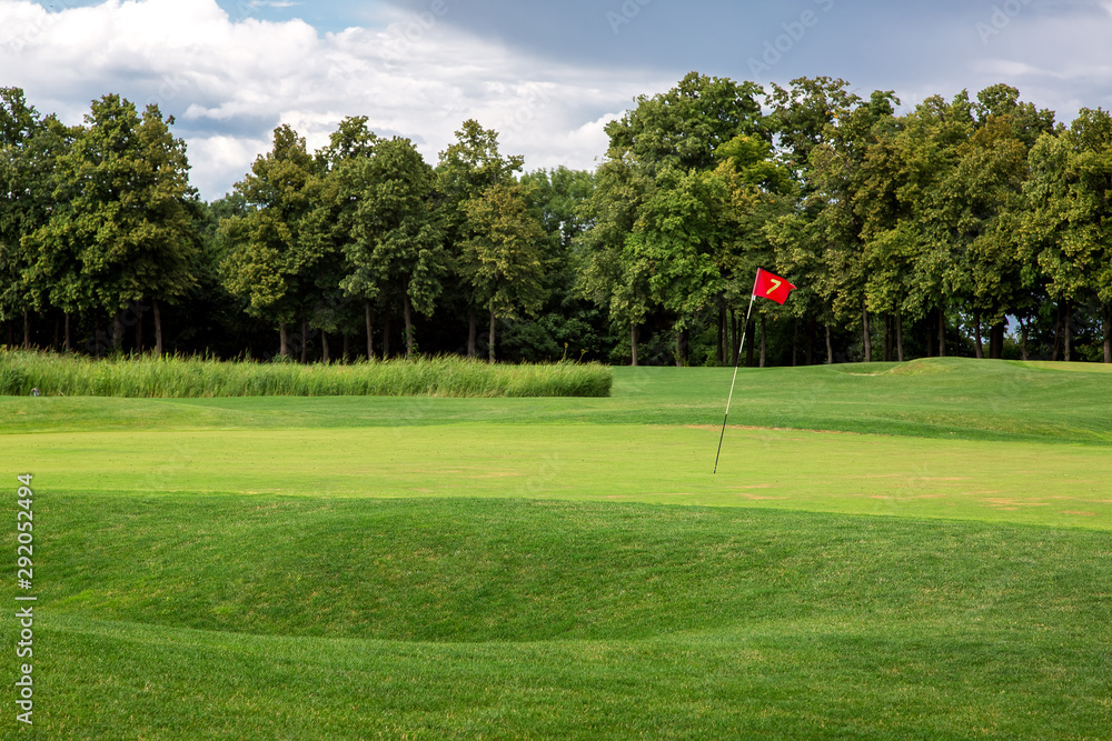 andscape of a golf course with a green lawn and a hilly surface of the playing space with a hole marked with a red flag, in the background cane and trees on a sunny summer day. andscape of a golf course with a green lawn and a hilly surface of the playing space with a hole marked with a red flag, in the background cane and trees on a sunny summer day.