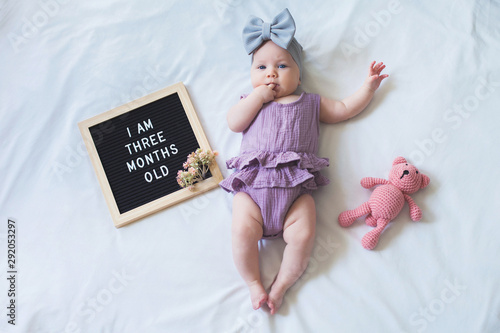 Shot from overhead of three months old baby girl laying down on white background with letter board and teddy bear.