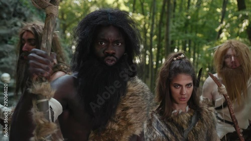 Wild primeval people of prehistoric multi-race tribe growling beating with sticks looking at camera. Hunter-gatherers nomadic family in the woods. photo