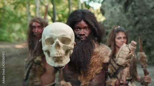 Savage tribe of primitive hunter-gatherers showing human skull at camera. Dreadful survival of prehistoric people. Evolution concept.