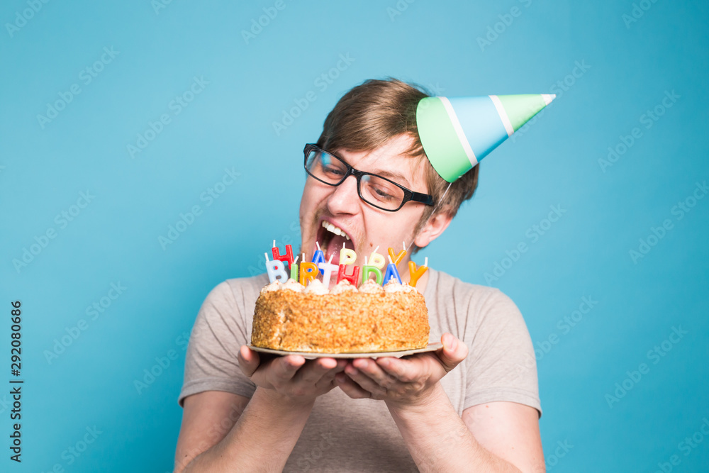 Funny male in paper congratulatory hat trying to bite off a cake with a happy birthday candles stand on a blue background.