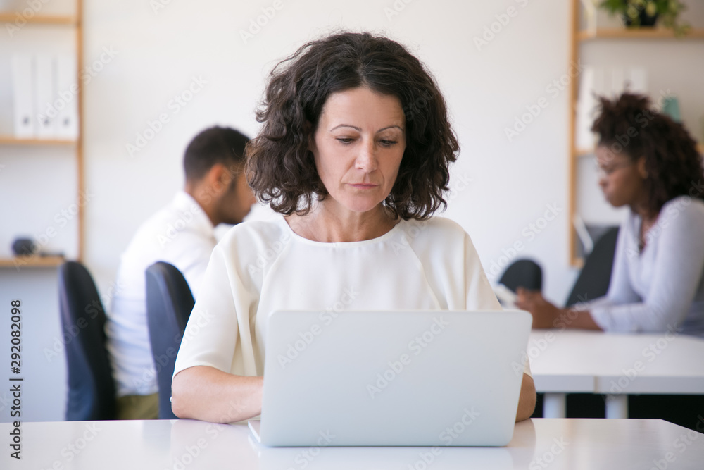Focused female employee working on laptop in office. Her diverse colleagues meeting at table and talking in background. Workspace concept Focused female employee working on laptop in office. Her diverse colleagues meeting at table and talking in background. Workspace concept