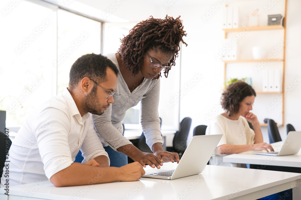 Fototapeta premium Instructor helping intern to start working laptop. Man and woman in casual sitting and standing at desk and using laptop. Internship concept