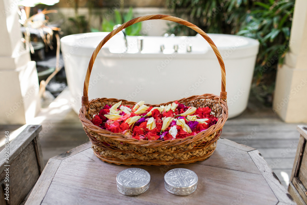 Wooden wicker basket with red and white flower petals in front of white stone empty bath over green tropical nature background. Spa relaxation concept.