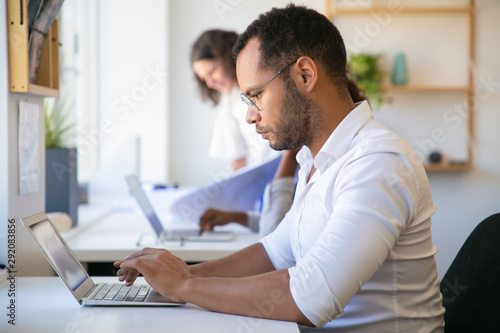 Focused male employee working on project in office. Man and women in casual sitting and standing at table and using laptops. Diverse staff concept