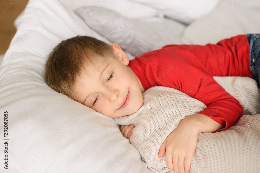 cute blond boy in red jersey sleeps among white pillows, blankets and hugs cute blond boy in red jersey sleeps among white pillows, blankets and hugs