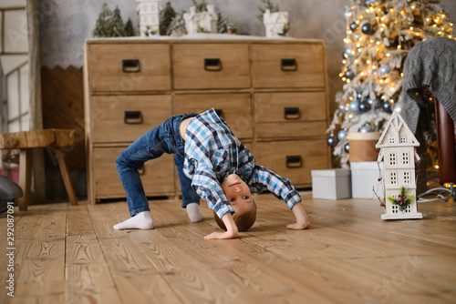boy stands in an acrobatic pose on  wooden floor, against background of Christmas tree, glowing with lights and bokeh