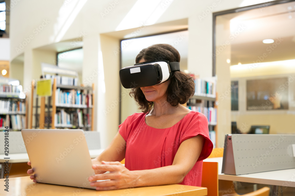 Adult female student using VR simulator while doing research in library.  Woman wearing virtual reality glasses, sitting at desk with laptop. Working  on research concept foto de Stock | Adobe Stock
