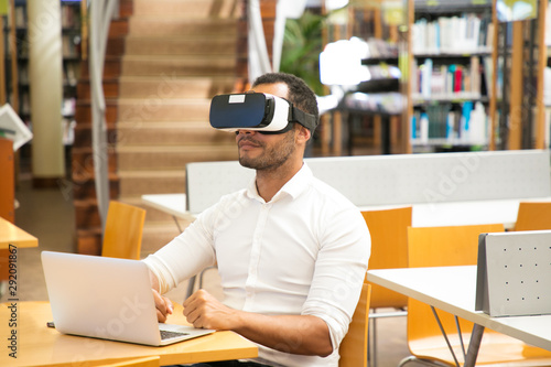 Male student using VR headset during work in library. Latin man wearing virtual reality glasses, sitting at desk with laptop. Education concept