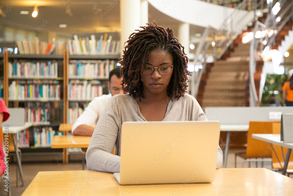 Serious African American student working on research in library. Students sitting at desks and using laptops in computer class. Technology or education concept Serious African American student working on research in library. Students sitting at desks and using laptops in computer class. Technology or education concept
