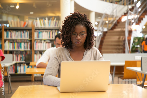 Serious African American student working on research in library. Students sitting at desks and using laptops in computer class. Technology or education concept