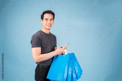 Handsome Asian man holding smartphone over blue wall background. holding shopping bags. 