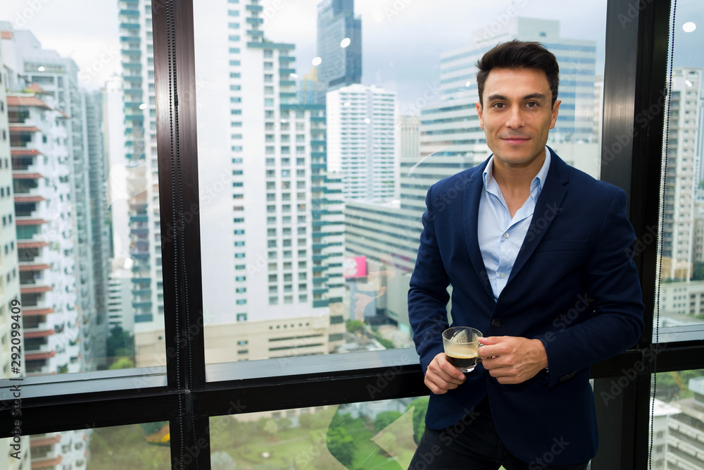 Adult handsome businessman who wears a formal suit is standing relaxed during a break from work, complete with a cup of coffee in a modern office surrounded by glass and have high-rise building views. Adult handsome businessman who wears a formal suit is standing relaxed during a break from work, complete with a cup of coffee in a modern office surrounded by glass and have high-rise building views.
