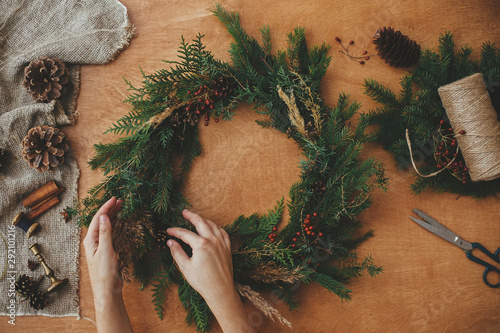 Quadro em tela Hands holding christmas wreath with fir branches, berries, pine cones, and thread, scissors on rural wooden table