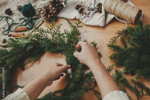 Hands holding fir branches, and pine cones, thread, berries, scissors on wooden table. Christmas wreath workshop. Authentic stylish still life. Making rustic Christmas wreath.