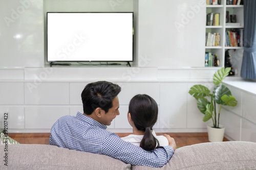Back view of young family, man and woman watching TV together in living room, TV on white background