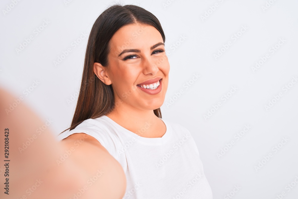 Beautiful young woman taking a selfie photo using smartphone over white background with a happy face standing and smiling with a confident smile showing teeth Beautiful young woman taking a selfie photo using smartphone over white background with a happy face standing and smiling with a confident smile showing teeth