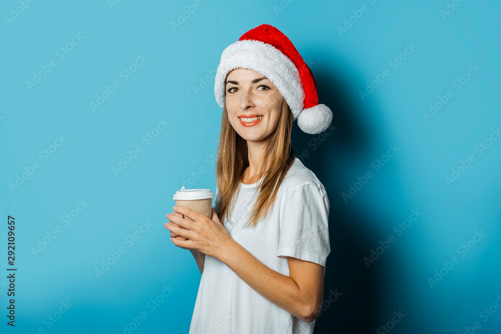 Beautiful Young woman with a smile in a hat of Santa Claus holds a cup of coffee on a blue background. Concept idea for New Year and Christmas, Christmas presents Beautiful Young woman with a smile in a hat of Santa Claus holds a cup of coffee on a blue background. Concept idea for New Year and Christmas, Christmas presents