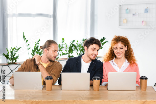 three smiling friends sitting at table and using laptops in office