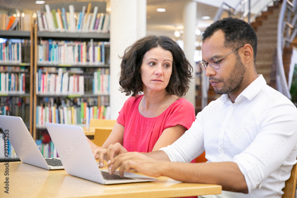 Two adult college students working in library computer class. Man and woman in casual sitting at desk with laptops, typing and talking. Education and teamwork concept Two adult college students working in library computer class. Man and woman in casual sitting at desk with laptops, typing and talking. Education and teamwork concept