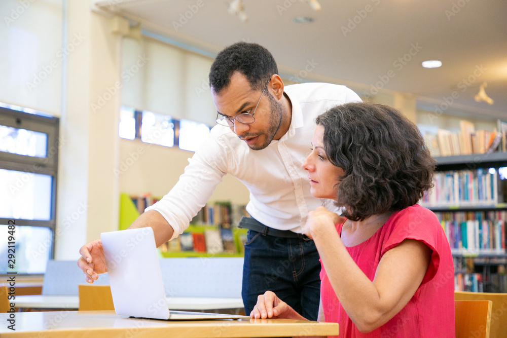 Instructor checking student work in library. Man and woman in casual sitting and standing at desk, using laptop, looking at monitor and talking. Training or course concept Instructor checking student work in library. Man and woman in casual sitting and standing at desk, using laptop, looking at monitor and talking. Training or course concept