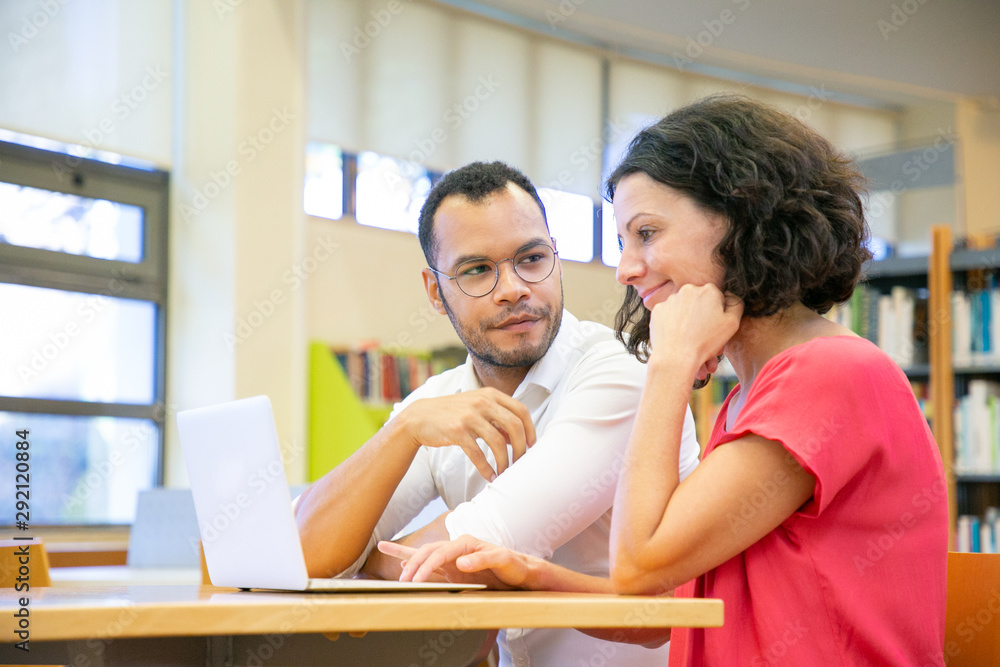 Two colleagues working on presentation in library. Man and woman in casual sitting at desk, using laptop and talking. Bookshelves in background. Exploring or research concept Two colleagues working on presentation in library. Man and woman in casual sitting at desk, using laptop and talking. Bookshelves in background. Exploring or research concept