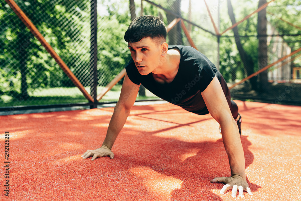 Young athletic muscular man doing push-ups on workout ground. Caucasian fitness male doing workout exercises on sunset light background. People and sport concept