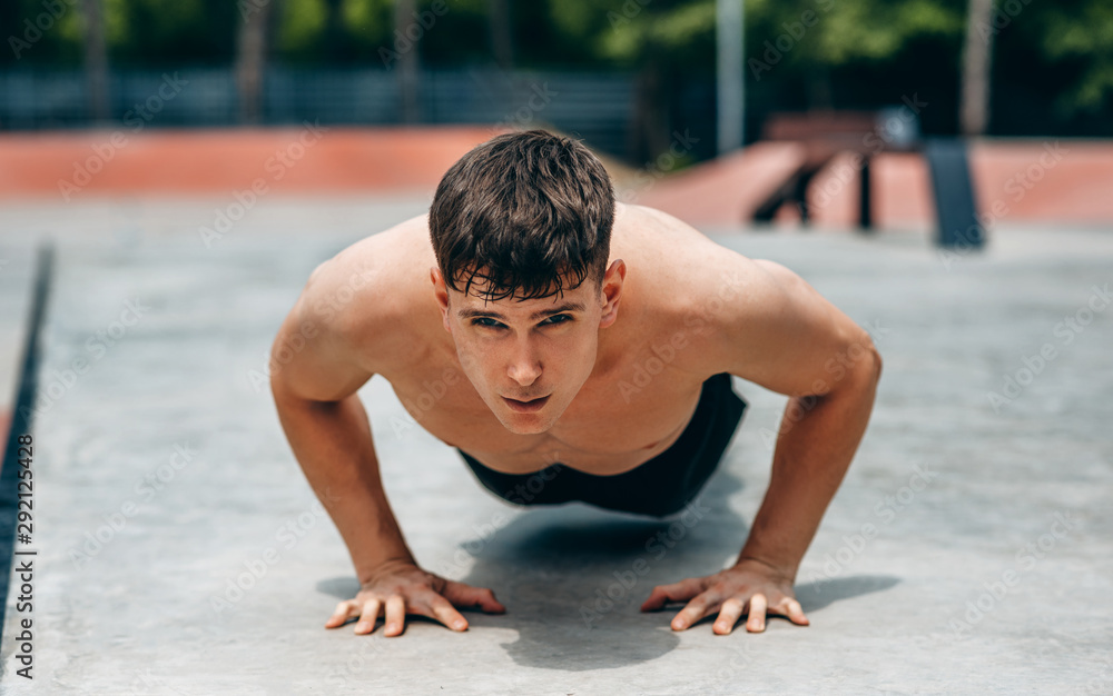 Fototapeta premium Image of young athletic muscular man doing push-ups on workout ground. Caucasian fitness male doing workout exercises on sunset light background. People and sport concept