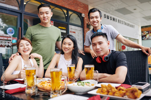 Portrait of Asian young friends smiling at camera while sitting at the table with glasses of beer during their meeting at the restaurant