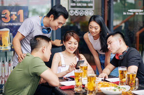 Happy Asian woman sitting with mobile phone and showing to her friends funny photos on it during their meeting in cafe © DragonImages