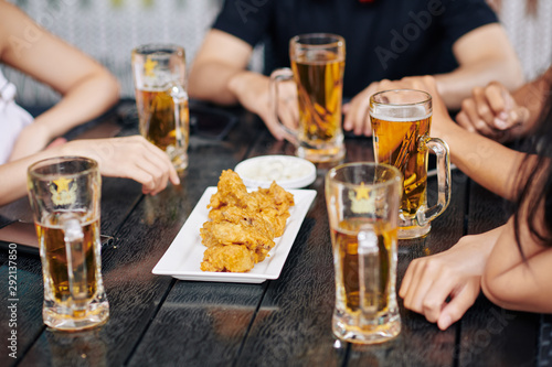 Close-up of friends sitting at wooden table and drinking beer from the glasses and eating fried chicken in cafe