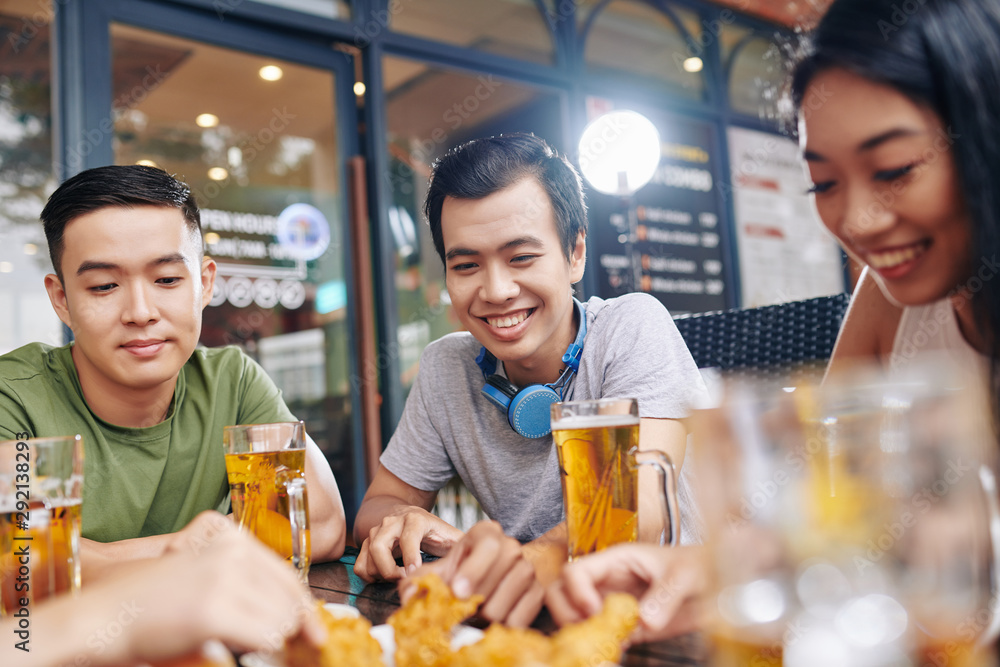 Group of Asian young people sitting at the table and laughing while eating chicken and drinking beer in cafe