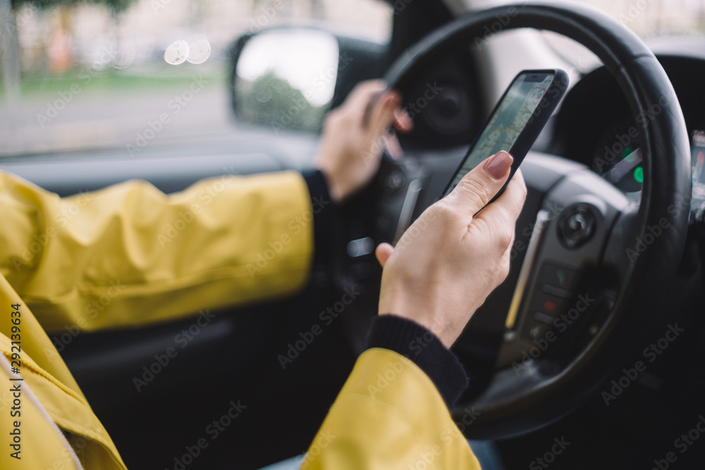 Close up photo of unrecognizable girl, who drive car and hold smartphone in the hand. Female use navigation maps while driving. Tourist concept. Selective focus on arm. Yellow jacket.