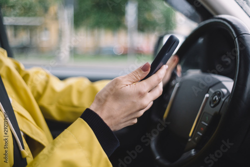 Close up photo of unrecognizable girl, who drive car and hold smartphone in the hand. Female use cellphone while driving. Tourist concept. Selective focus on arm. Yellow jacket. Blurred background.