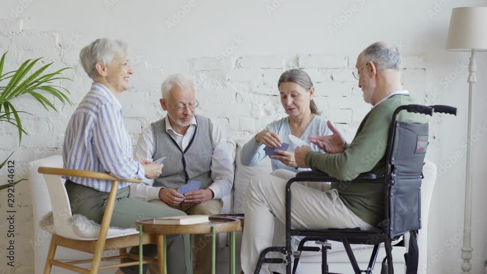 Group of four elderly people, including disabled man on wheelchair talking while playing cards sitting together in nursing home Group of four elderly people, including disabled man on wheelchair talking while playing cards sitting together in nursing home