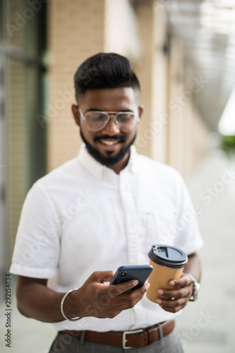 Young attractive man dressed in trendy outfit monitoring information from social networks via cellphone connecting to 4G internet while enjoying coffee to go strolling on megalopolis street