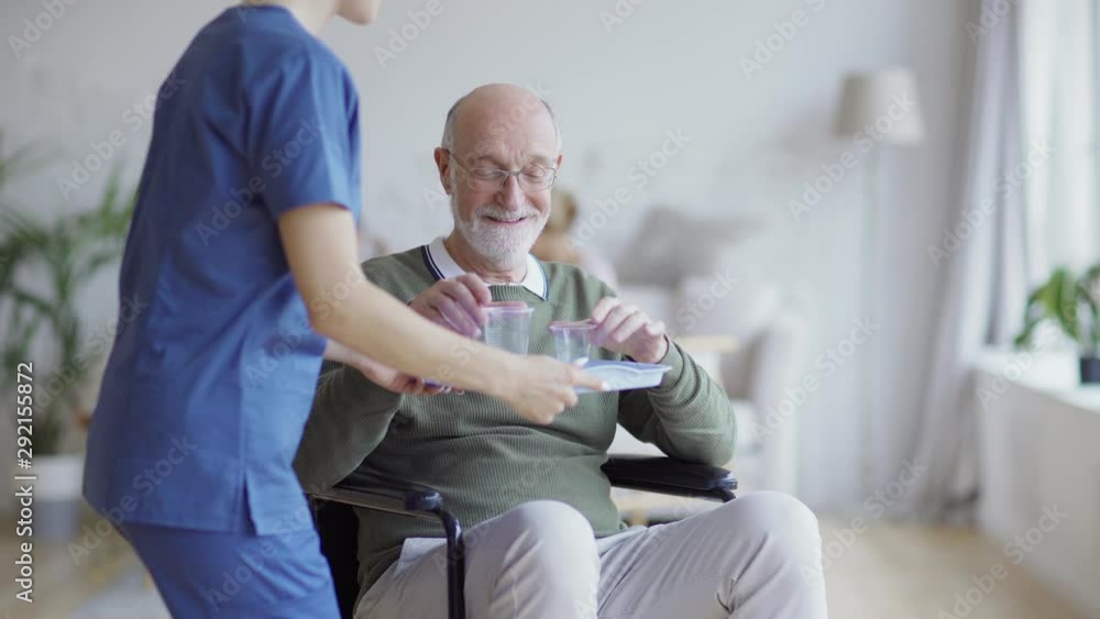 Disabled elderly man in eyeglasses sitting in wheelchair and looking away thoughtfully. Senior patient smiling and looking at camera when female nurse bringing him vitamin pills and glass of water Disabled elderly man in eyeglasses sitting in wheelchair and looking away thoughtfully. Senior patient smiling and looking at camera when female nurse bringing him vitamin pills and glass of water