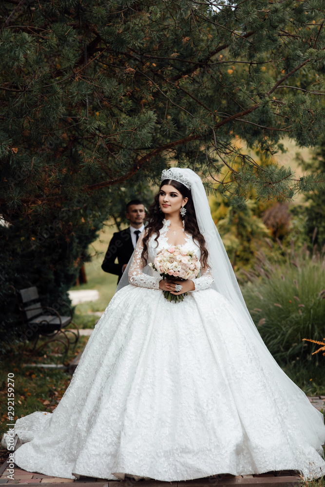 Handsome groom in black formal suit and his elegant bride in dress and veil with bouquet with beautiful hairdress. Pair embrace on a walk in the big green park. Beautiful wedding photosession.