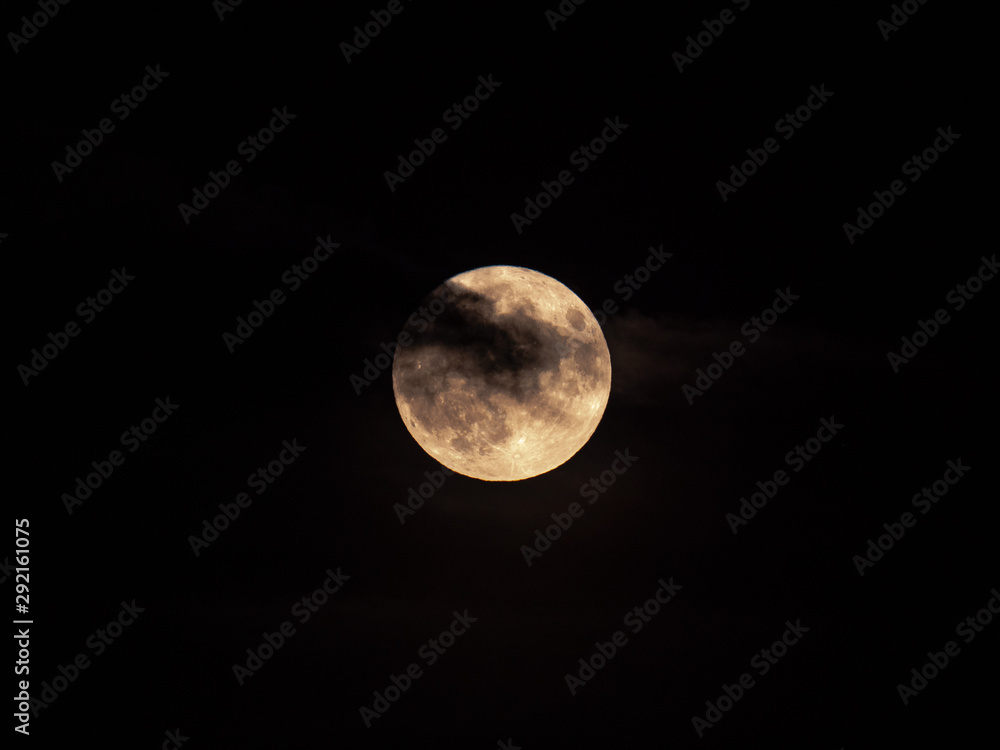 Fototapeta premium A closeup view of the micro harvest full moon with partial cloud cover and haze on Friday the 13th as seen in Chicago on a black night sky backdrop making for a creepy, spooky or scary scene.