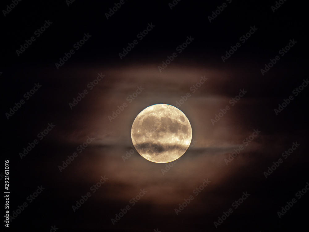 A closeup view of the micro harvest full moon with partial cloud cover and halo on Friday the 13th as seen in Chicago on a black night sky backdrop making for a creepy, spooky or scary scene.