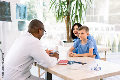 cheerful caucasian mother and her son in doctor's office with african doctor, sitting at the table. Healthcare and pediatrics concept