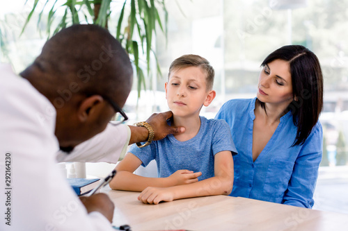African-American male doctor in his modern office clinic examining throat of little boy with pretty mother watching.
