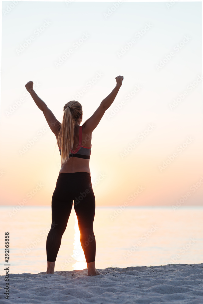 Young sporty woman standing by the sea with her arms raised in the air in sunset