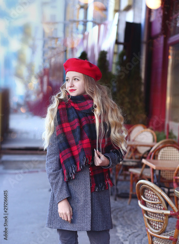 girl with a smile in a gray coat and a red beret with long curls stands thoughtfully near a cafe on a background of a red facade photo