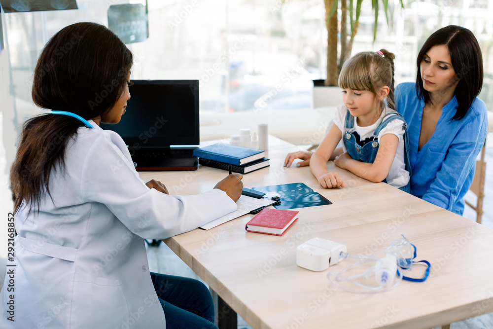 Side view of African-American female doctor discussing over x-ray report with Caucasian mother and daughter in the modern office at hospital. Laptop, nebulizer, x-ray on the table Side view of African-American female doctor discussing over x-ray report with Caucasian mother and daughter in the modern office at hospital. Laptop, nebulizer, x-ray on the table